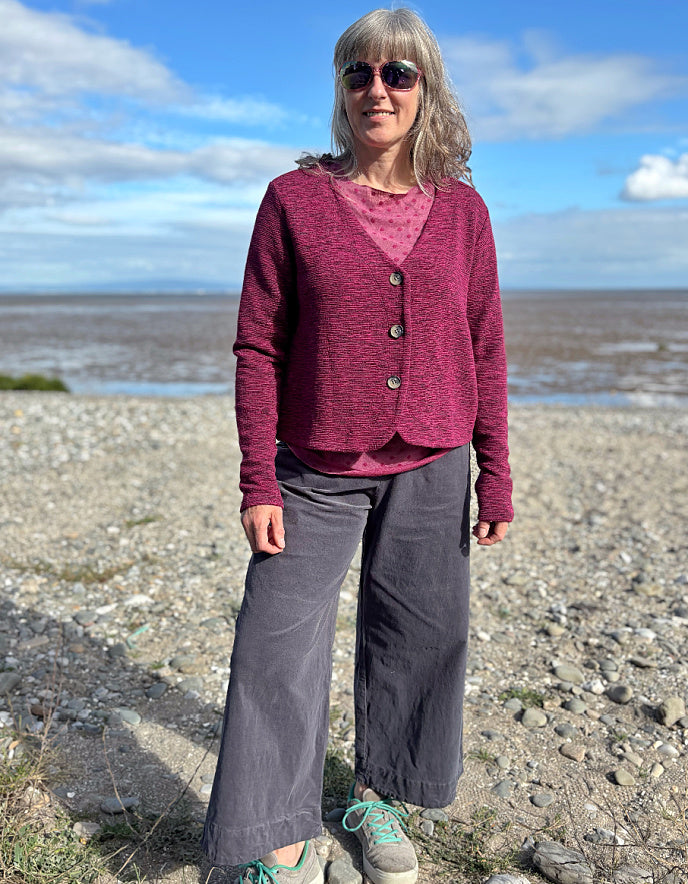 Woman standing on a pebbly beach wearing a purple cardigan and gray pants.