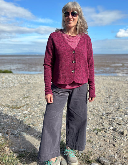 Woman standing on a pebbly beach wearing a purple cardigan and gray pants.