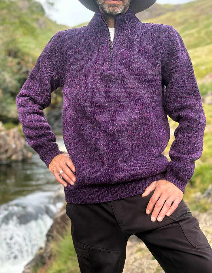 A man wearing a textured purple quarter-zip knit sweater and black pants, standing outdoors near a stream with green hills in the background.