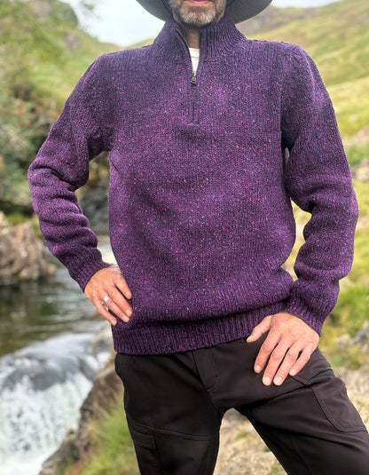 A man wearing a textured purple quarter-zip knit sweater and black pants, standing outdoors near a stream with green hills in the background.