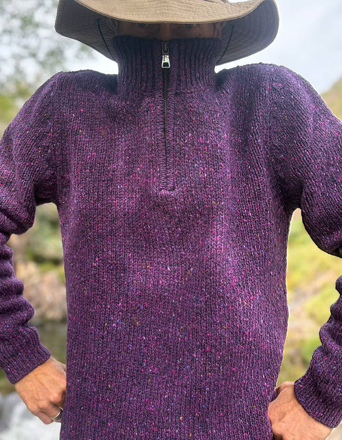 A person wearing a purple half-zip knit sweater with flecks of pink and blue yarn, paired with a wide-brimmed brown hat, photographed outdoors with a blurred natural background.
