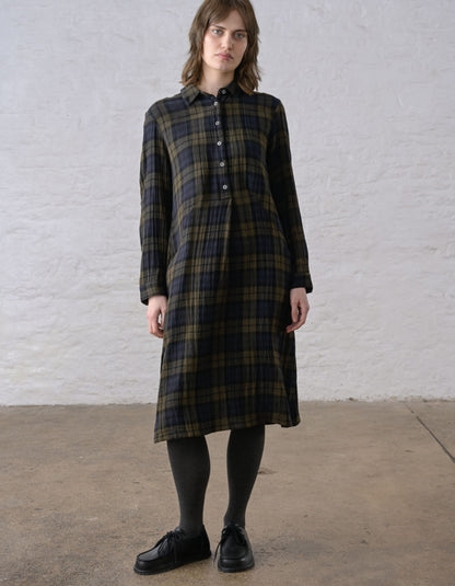 A woman stands against a white textured wall wearing a long-sleeve plaid shirt dress in dark green, black, and brown tones, paired with black tights and black shoes.
