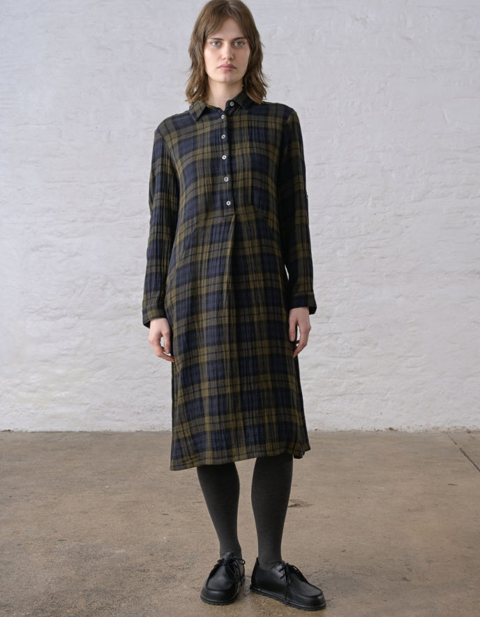 A woman stands against a white textured wall wearing a long-sleeve button-front plaid dress in dark green, black, and brown tones, paired with black tights and black shoes.