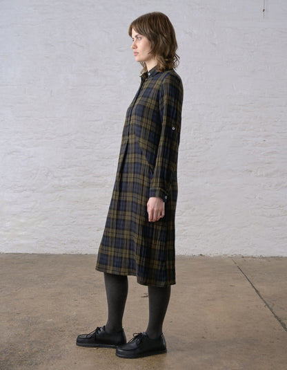 A woman stands in profile wearing a long plaid shirt dress with a dark navy and olive green pattern, paired with black tights and black shoes, against a white brick wall.