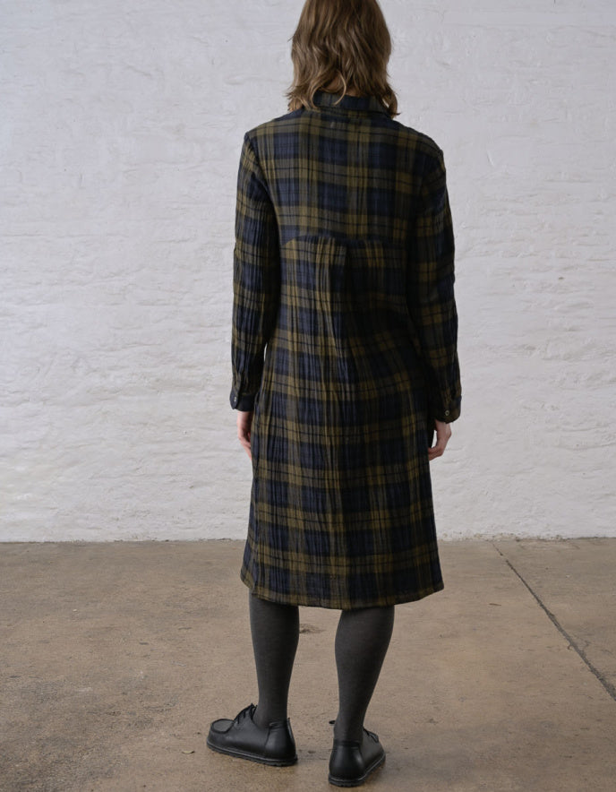 A person shown from the back wearing a long plaid coat in dark green, black, and tan, standing against a white textured wall on a concrete floor.
