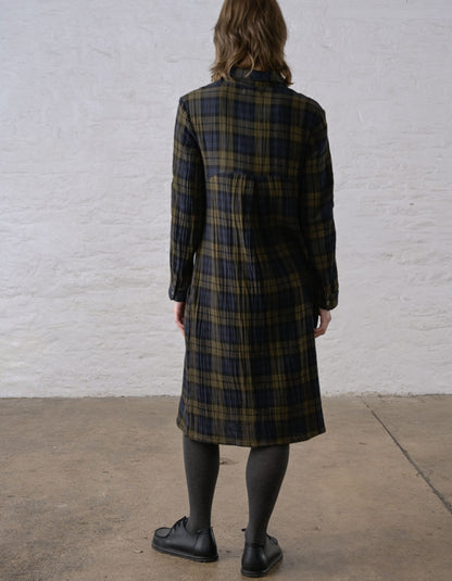 A person shown from the back wearing a long plaid coat in dark green, black, and tan, standing against a white textured wall on a concrete floor.