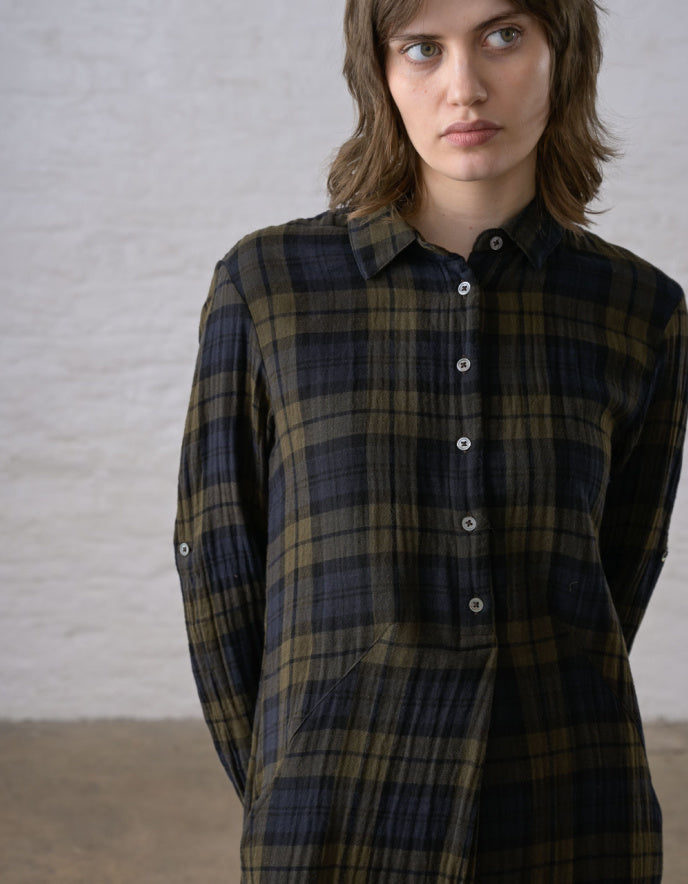 A woman wearing a dark plaid button-up shirt with olive green and navy stripes, photographed against a light textured wall.