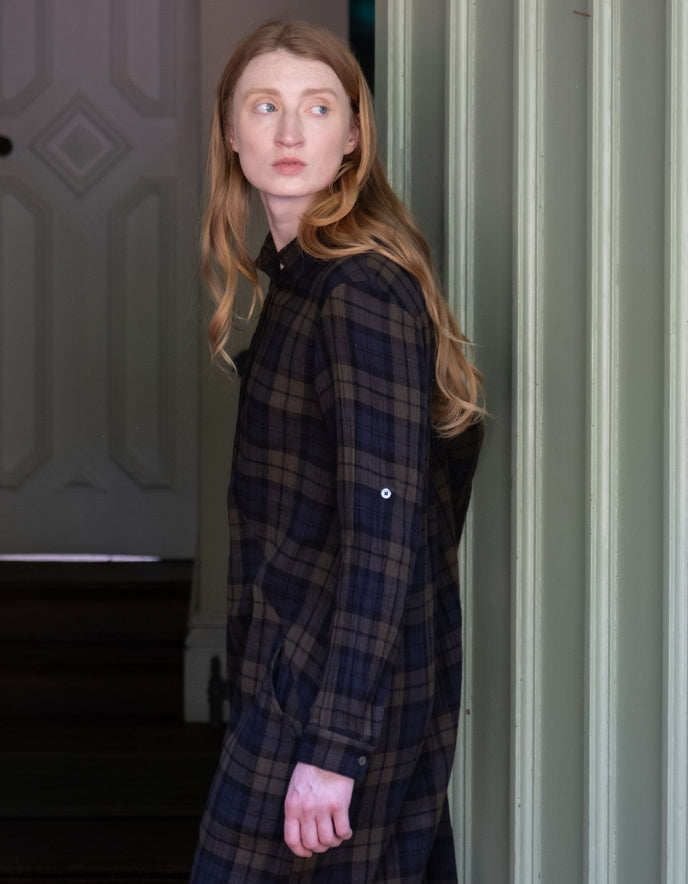 A woman with long light brown hair wearing a dark navy and brown plaid shirt jacket, standing in a doorway.