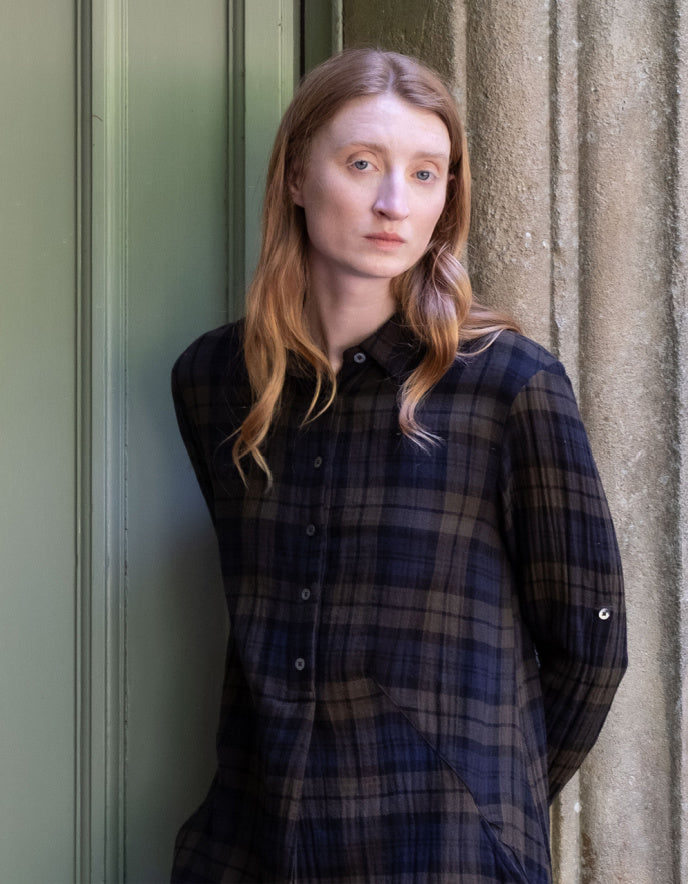 A woman with long light brown hair wearing a dark navy and black plaid button-up shirt, leaning against a textured stone wall next to a green door.