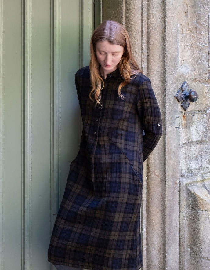 A woman with long light brown hair stands leaning against a stone wall next to a muted green door, wearing a long-sleeved navy and brown plaid button-up shirt dress.