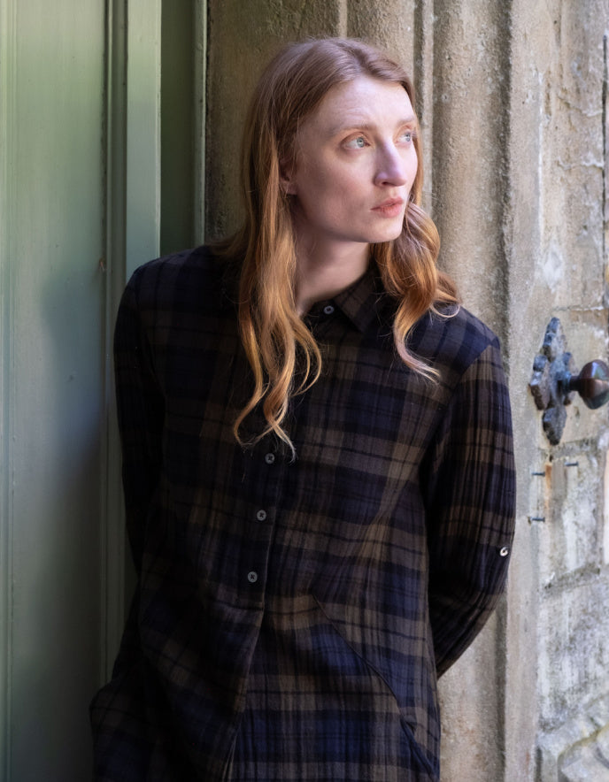 A woman with long light brown hair wearing a dark navy and brown plaid button-up shirt, leaning against a rustic stone wall next to a green door.