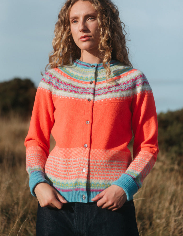 A woman wearing a bright coral-orange knitted cardigan with multicolored Fair Isle patterns across the yoke, cuffs, and hem, standing outdoors in a field.