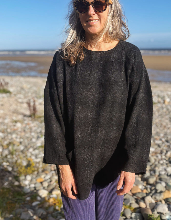 A woman wearing a loose, dark charcoal gray long-sleeve top and purple pants stands on a rocky beach with water and sky in the background.