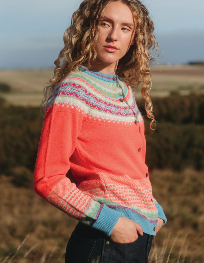 A woman wearing a coral pink button-up cardigan with a multicolor Fair Isle yoke and cuffs, standing outdoors in a grassy field.