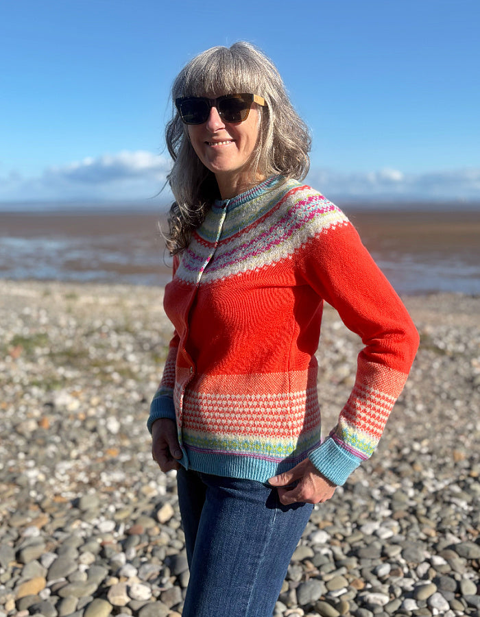 A woman wearing a colorful hand-knit zip-up cardigan sweater and blue jeans stands on a pebble beach with water and a blue sky in the background.