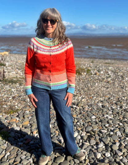 A woman wearing a colorful knit cardigan and blue jeans stands on a pebble beach with water and a blue sky in the background.