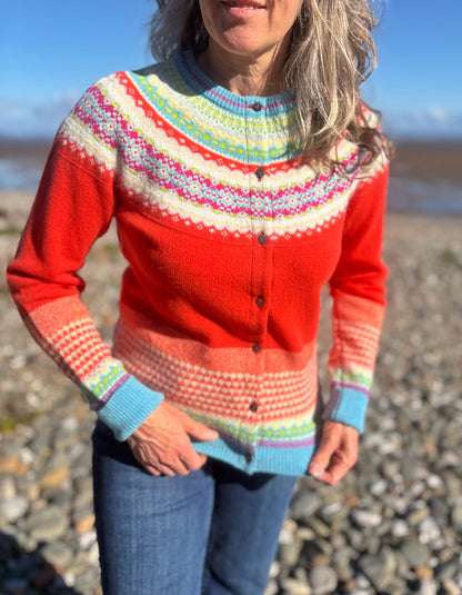 A woman wearing a bright red button-up cardigan sweater with multicolored Fair Isle patterns, standing on a rocky beach.