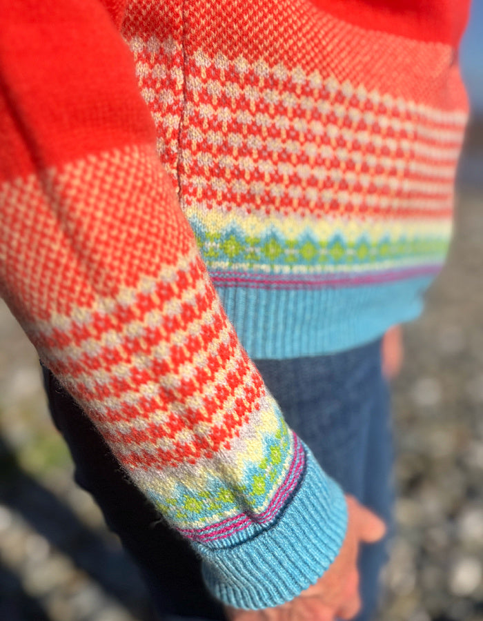 Close-up of a person wearing a colorful knit sweater with a red and cream patterned body, a light blue ribbed hem and cuff, and a multicolor geometric stripe detail, paired with dark blue jeans.