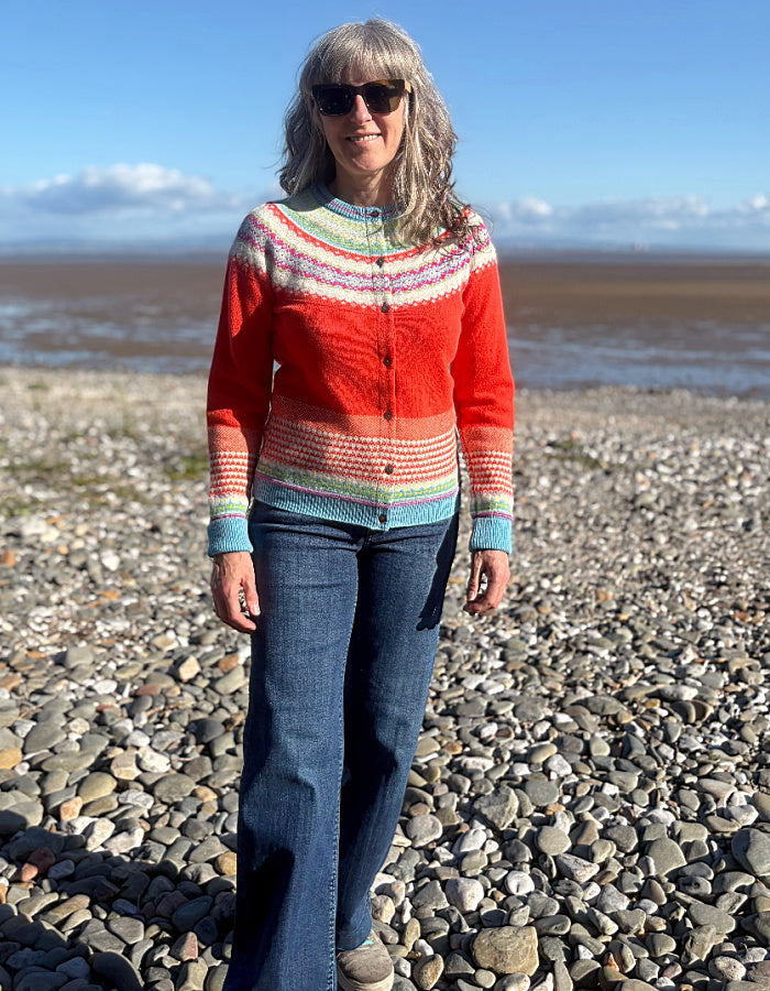 A woman wearing a colorful hand-knit cardigan and blue jeans stands on a pebble beach with water and a blue sky in the background.