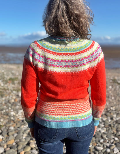 A woman viewed from behind wearing a bright red knit sweater with a multicolored Fair Isle patterned yoke and hem, standing on a pebble beach with water and sky in the background.