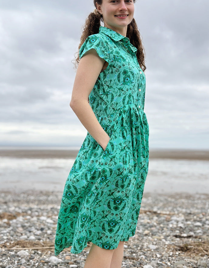 Woman wearing a green floral cotton shirt dress standing on a pebbly beach with cloudy skies.