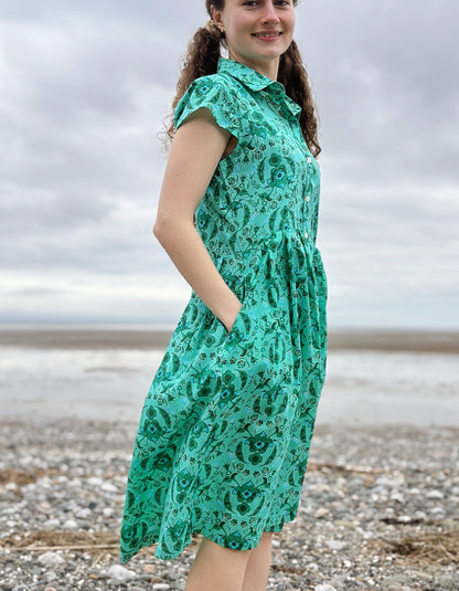 Woman wearing a green floral cotton shirt dress standing on a pebbly beach with cloudy skies.