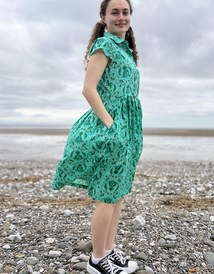 Woman wearing a green floral cotton shirt dress standing on a pebbly beach with cloudy skies.