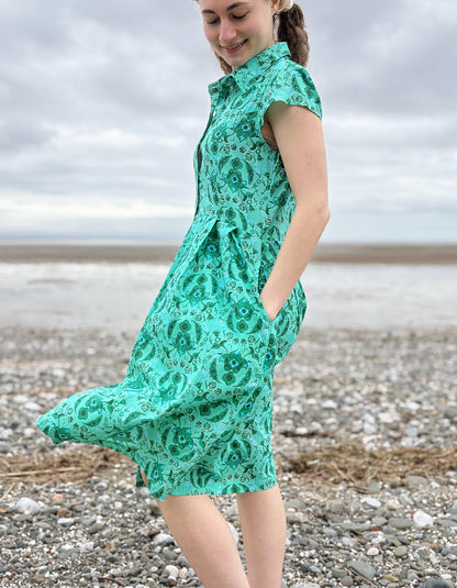 Woman wearing a green floral cotton shirt dress standing on a pebbly beach with cloudy skies.