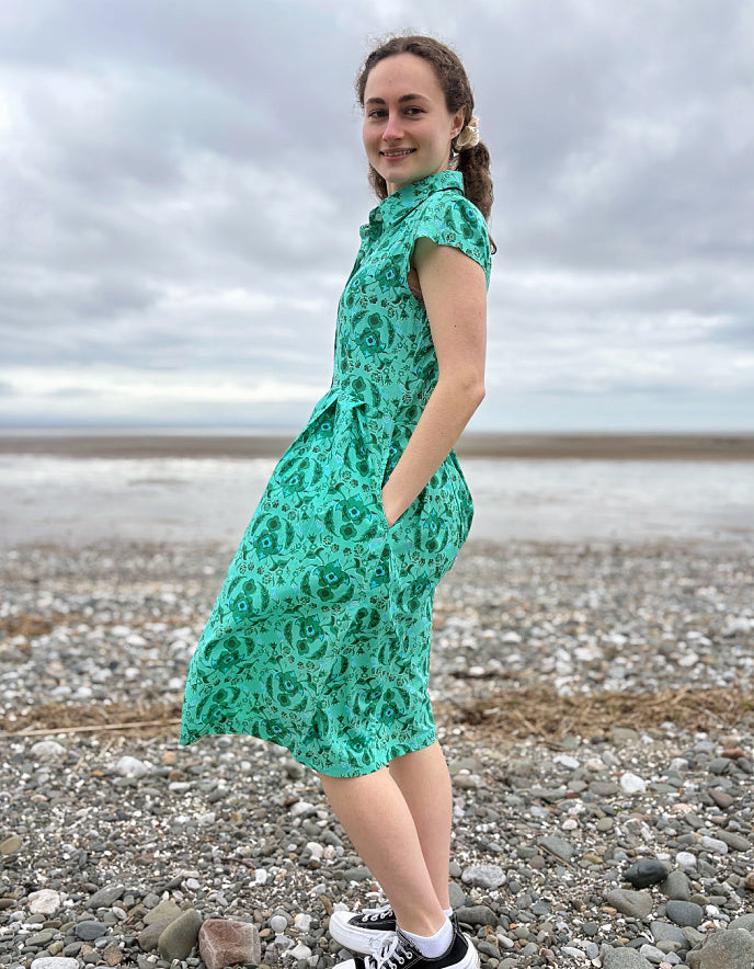 Woman wearing a green floral cotton shirt dress standing on a pebbly beach with cloudy skies.