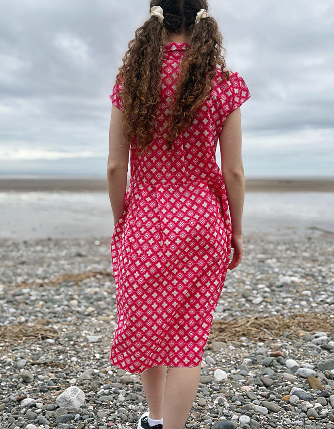 Woman wearing a pink cotton shirt dress with a geometric pattern on a pebbly beach.