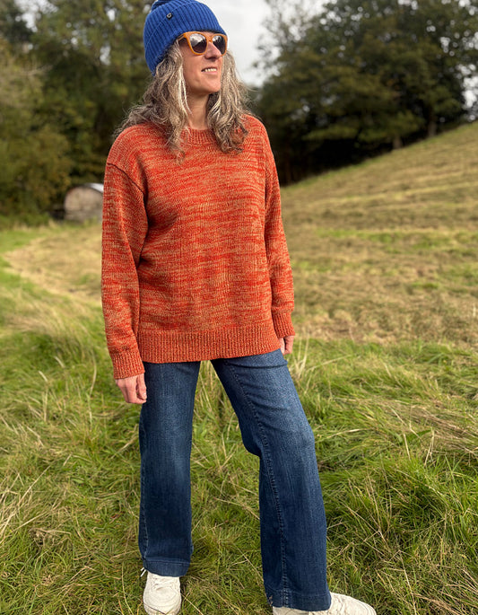 A woman standing outdoors in a grassy field wearing a marled orange knit sweater, blue jeans, a blue beanie, and white sneakers.