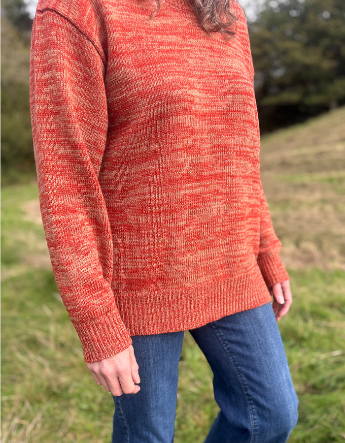 A person wearing a marled orange and beige knit sweater with blue jeans, standing outdoors in a grassy field.