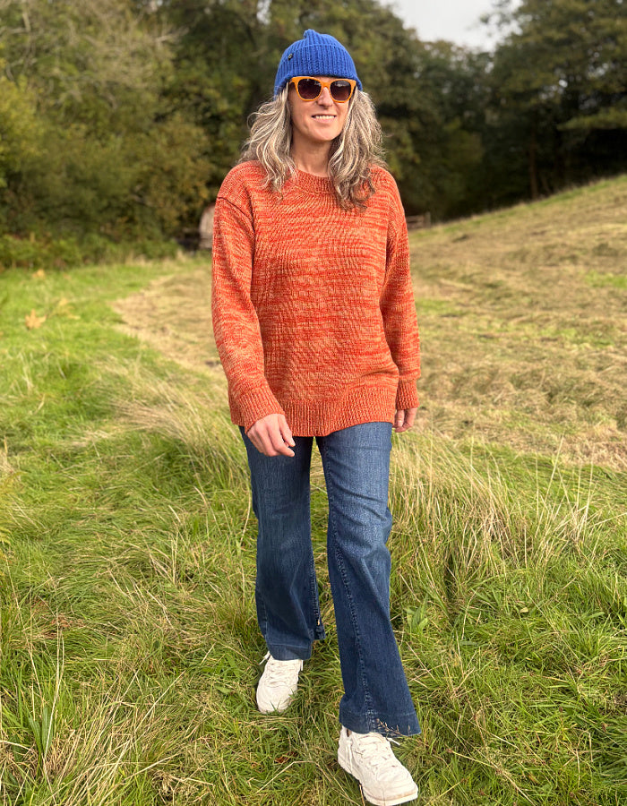 A woman standing in a grassy field wearing a textured orange knit sweater, blue jeans, white sneakers, a blue beanie, and sunglasses.