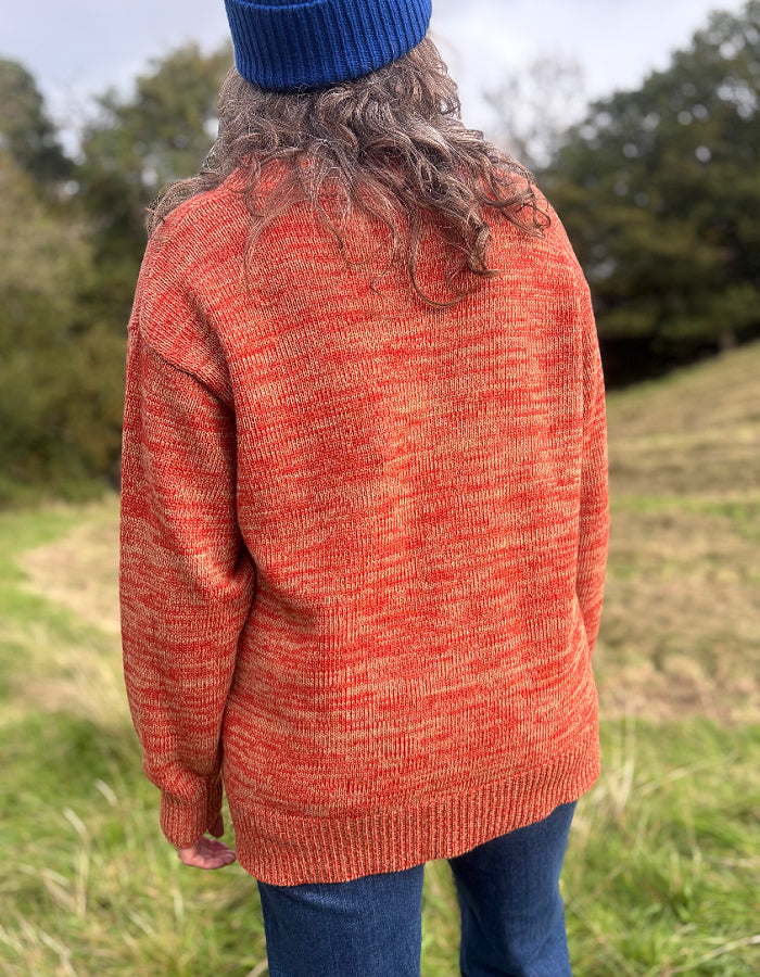 A person standing outdoors in a grassy field, viewed from behind, wearing a loose knit sweater in a heathered orange and beige colorway, blue jeans, and a blue beanie.