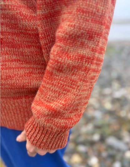 Close-up of a person wearing a marled knit sweater in orange and beige tones, with a blurred outdoor background.