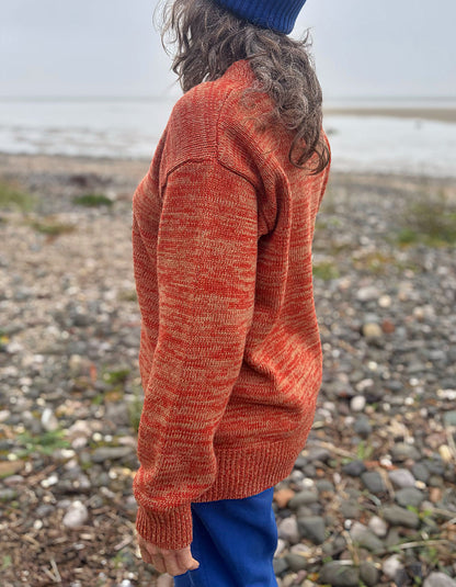 A person standing on a pebble beach wearing a marled orange and beige knit sweater, blue pants, and a blue beanie, shown from the back.