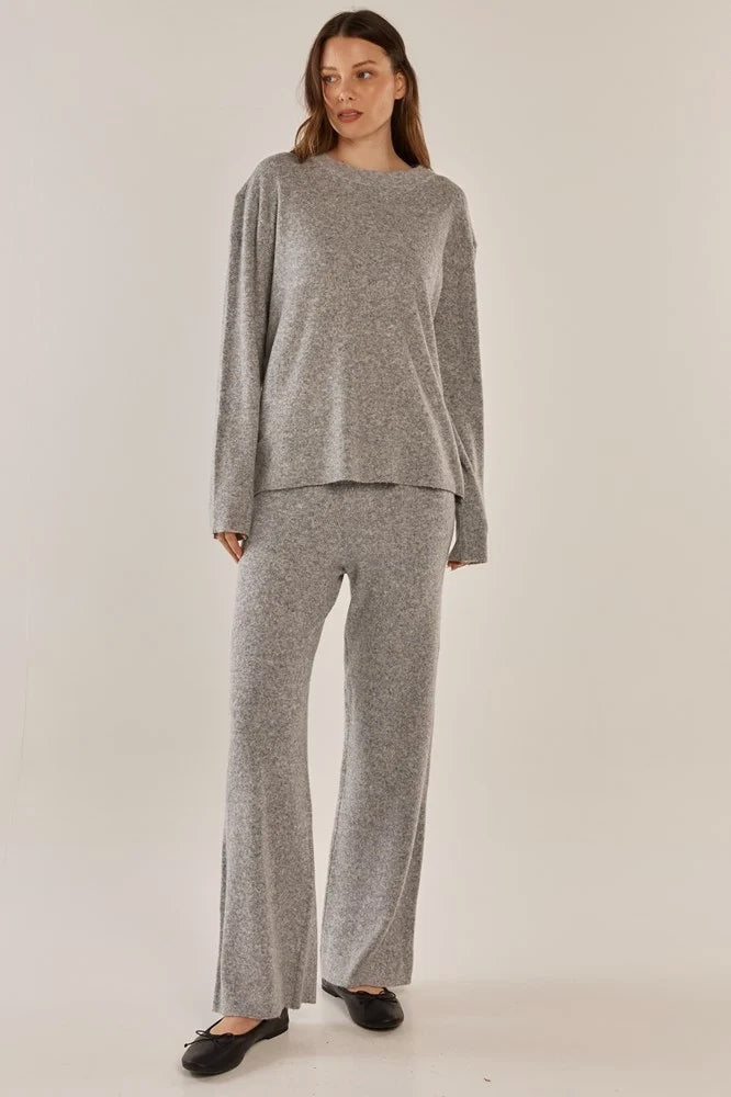 A woman modeling a matching light gray knit loungewear set consisting of a long-sleeve crewneck top and wide-leg pants, paired with black flats, against a plain light beige studio background.