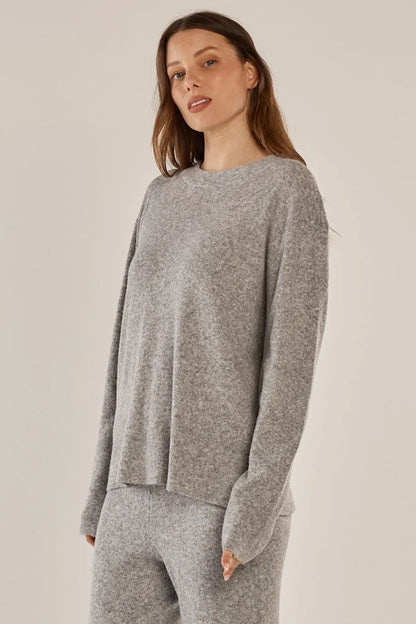 A woman modeling a light gray knit crewneck sweater and matching gray knit pants against a plain beige studio background.