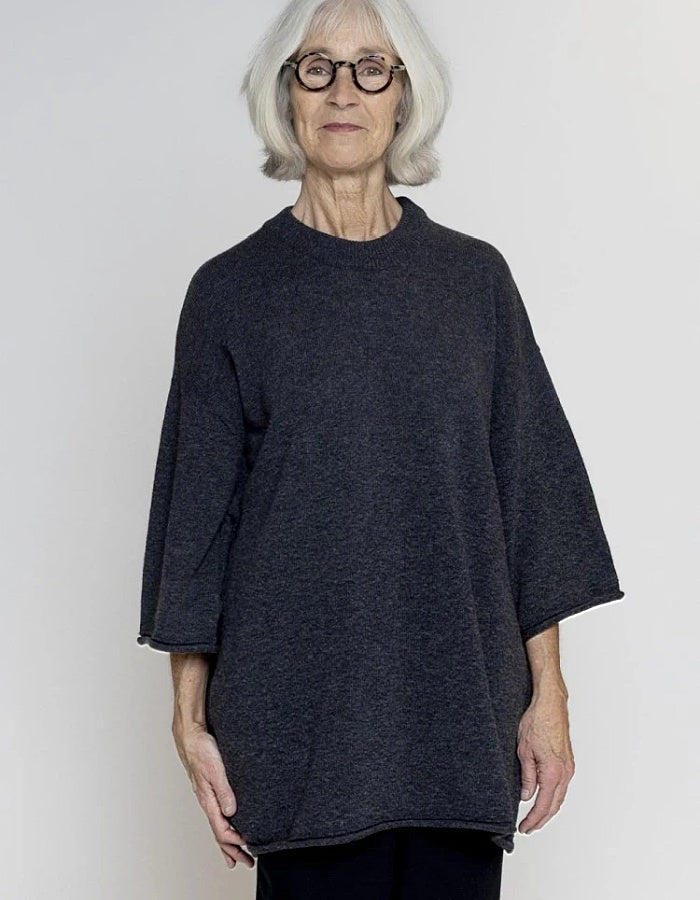 


An older woman with short grey hair is shown wearing a dark charcoal grey open-front cardigan or poncho-style top with wide three-quarter sleeves, against a plain light grey studio background.