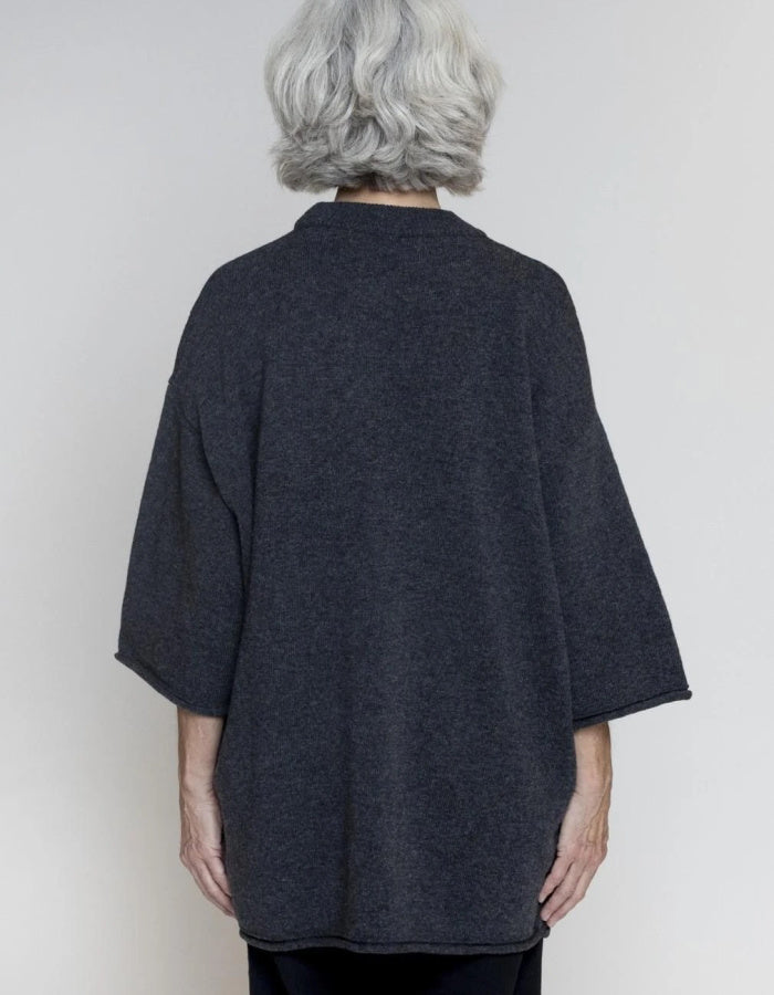 An older woman with short gray hair is shown from the back wearing a dark charcoal gray open-front cardigan or poncho-style top with wide three-quarter sleeves, against a plain light gray studio background.