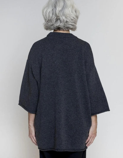 An older woman with short gray hair is shown from the back wearing a dark charcoal gray open-front cardigan or poncho-style top with wide three-quarter sleeves, against a plain light gray studio background.