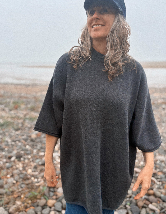 A woman with gray hair wearing a dark charcoal gray knit poncho sweater, blue jeans, and a navy cap, standing on a pebble beach.