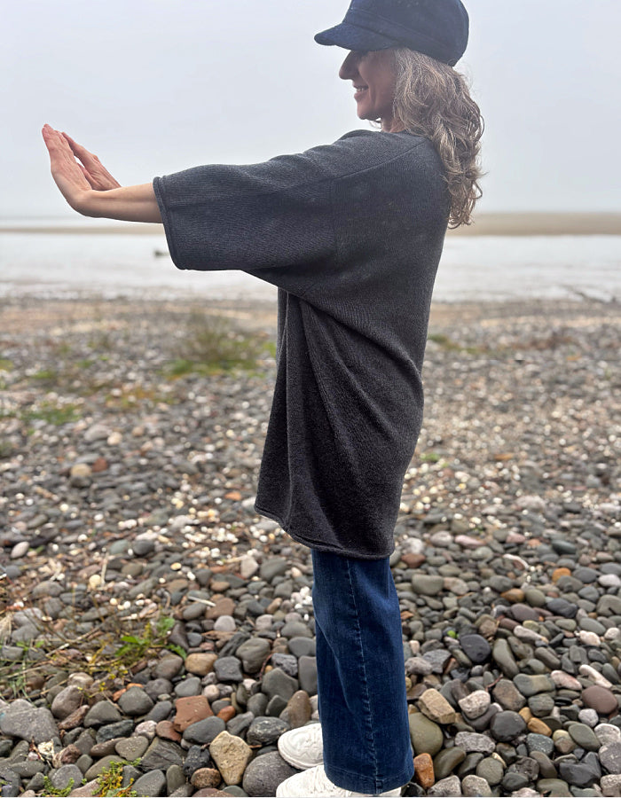 A woman standing on a pebble beach wearing a dark charcoal gray oversized t-shirt, blue jeans, a black cap, and white sneakers, with her arms outstretched.