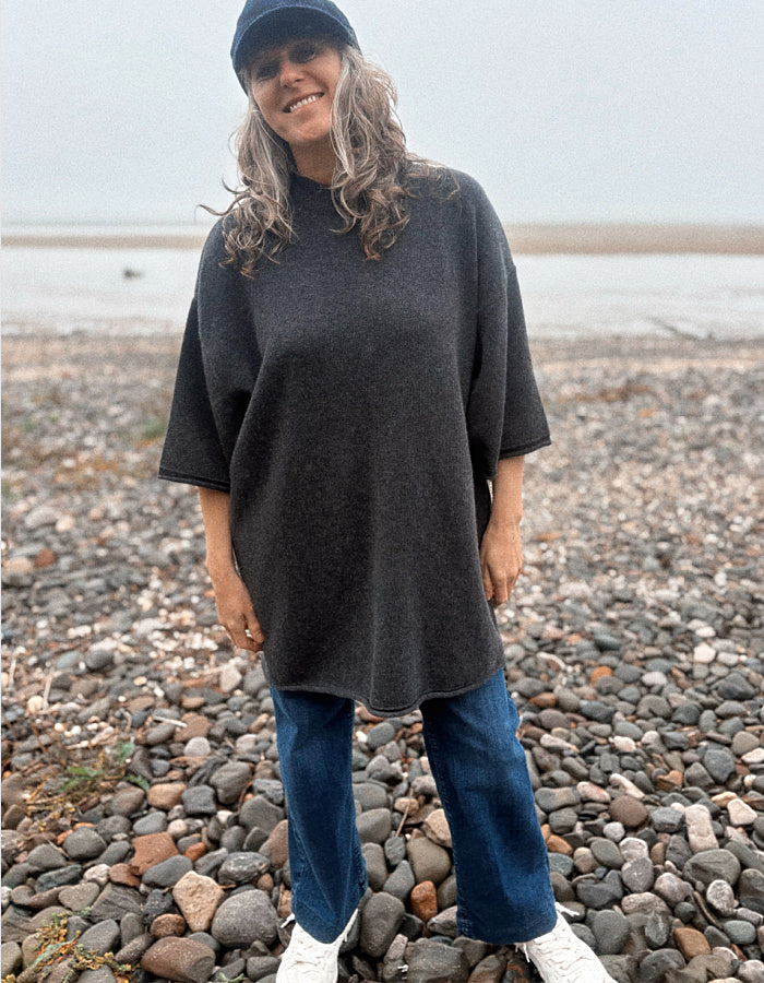 A woman wearing an oversized dark charcoal gray knit tunic sweater, blue jeans, and white sneakers stands on a pebble beach.