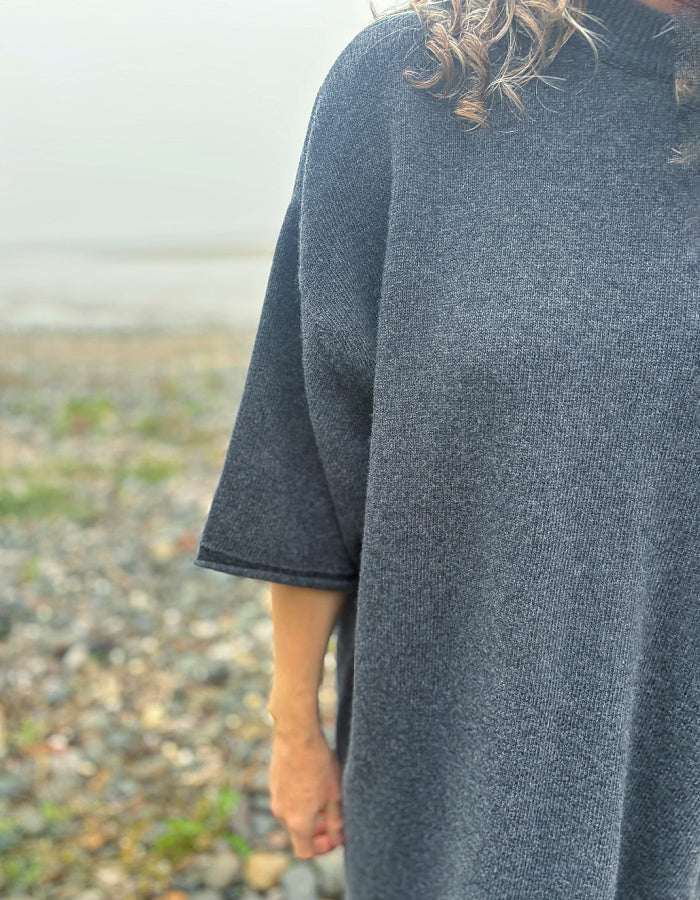 A close-up of a person wearing a dark charcoal gray knit poncho sweater outdoors on a pebble beach.