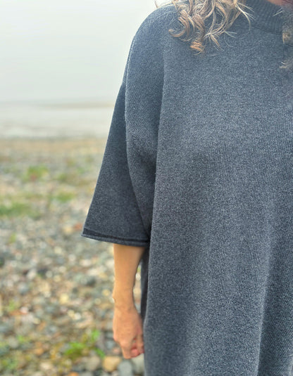 A close-up of a person wearing a dark charcoal gray knit poncho sweater outdoors on a pebble beach.
