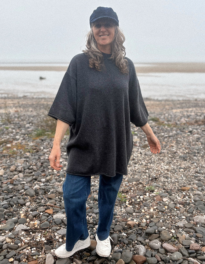A woman standing on a pebble beach wearing a dark charcoal gray oversized tunic top, blue jeans, white sneakers, and a dark baseball cap.