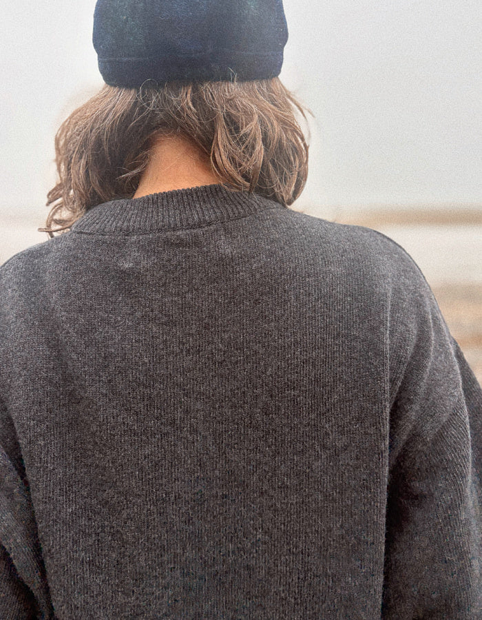 A person viewed from behind wearing a dark charcoal gray knit sweater and a black beanie, standing outdoors against a soft, hazy background.