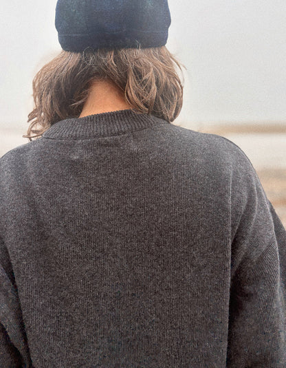 A person viewed from behind wearing a dark charcoal gray knit sweater and a black beanie, standing outdoors against a soft, hazy background.