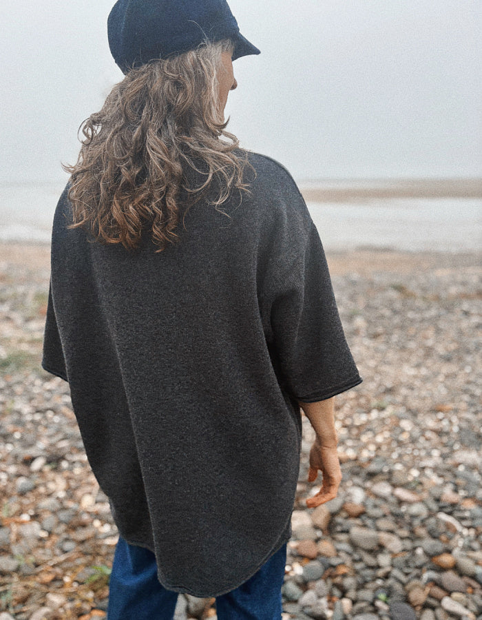 A person standing on a pebble beach, viewed from behind, wearing an oversized dark charcoal gray knit poncho-style top, blue jeans, and a black cap.